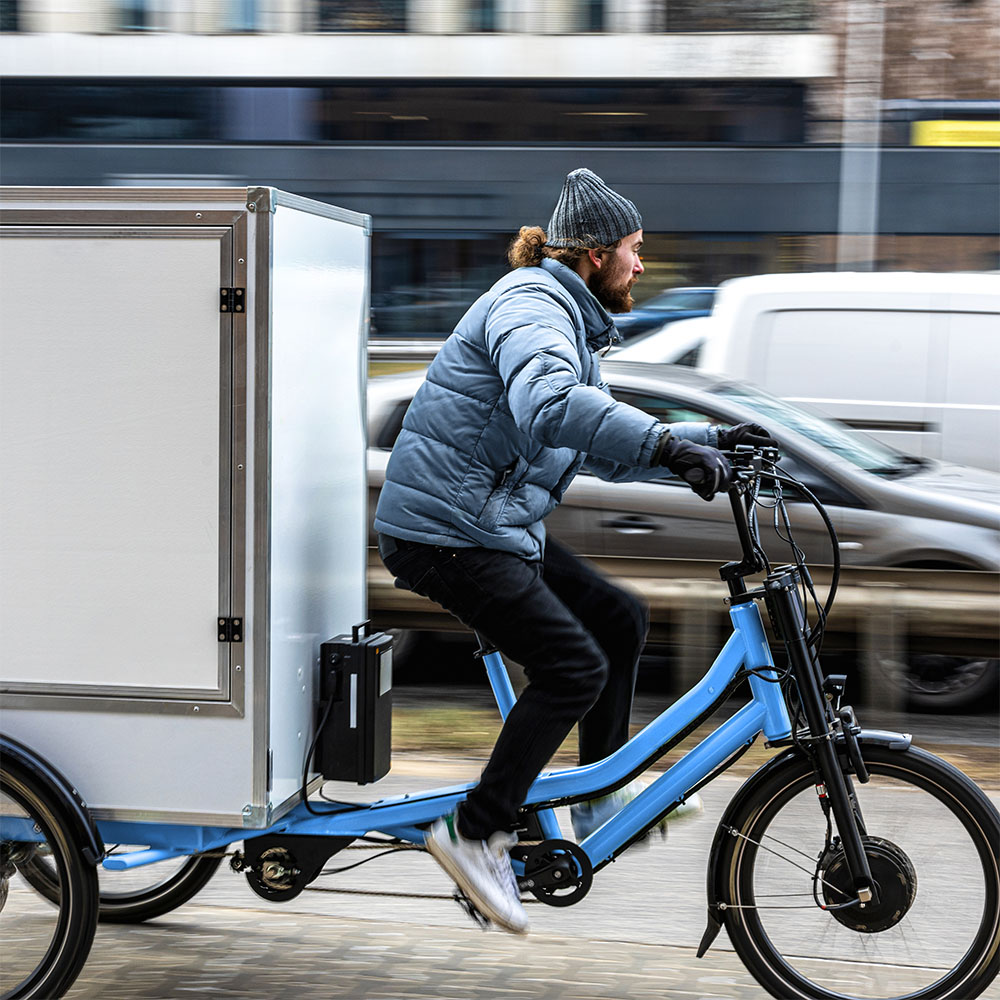 Junger Mann mit Bart auf einem Lastenrad durch die Innenstadt fahrend Junger Mann mit Bart auf einem Lastenrad durch die Innenstadt fahrend
